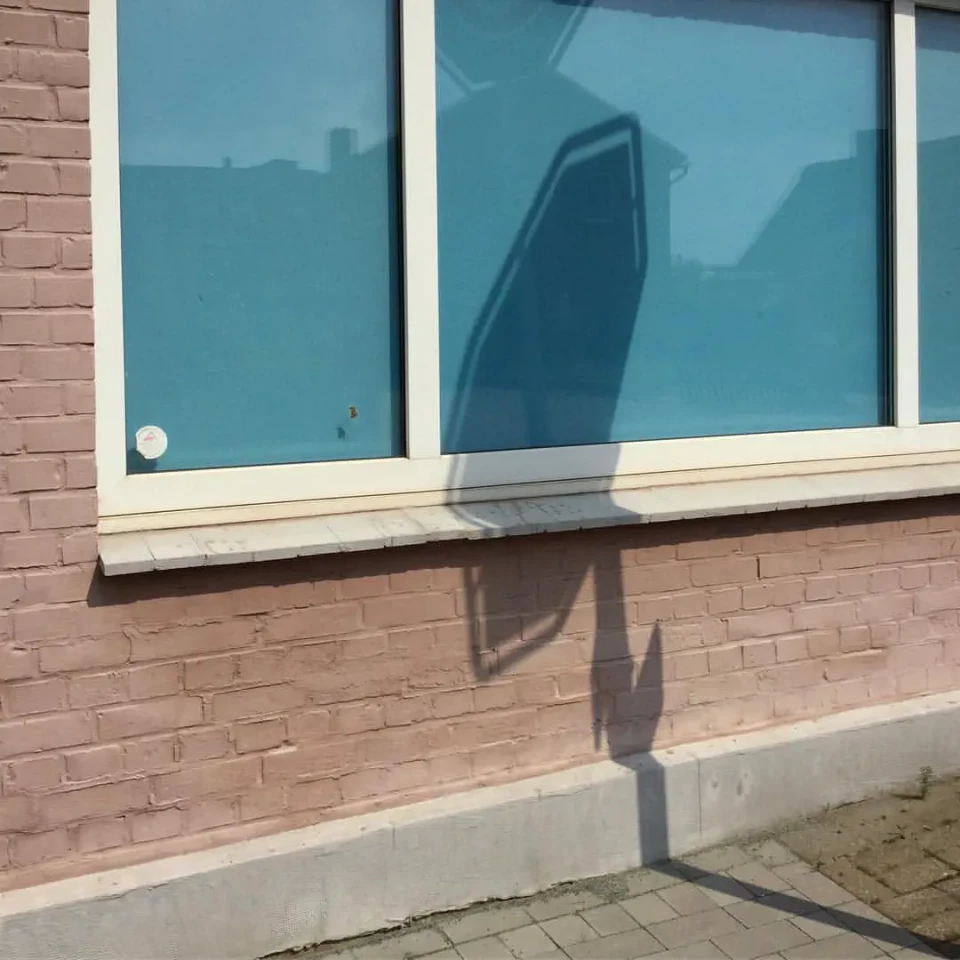 Das Foto zeigt eine rosa Hauswand mit hellblauem Fenster, in dem sich der Schatten von einem Verkehrsschild spiegelt