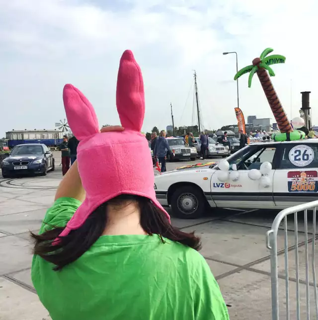 Das Bild zeigt eine Frau von hinten, die eine rosa Hasenmütze trägt. Sie ist auf einem Parkplatz, es sind Autos und eine Plastikpalme zu sehen