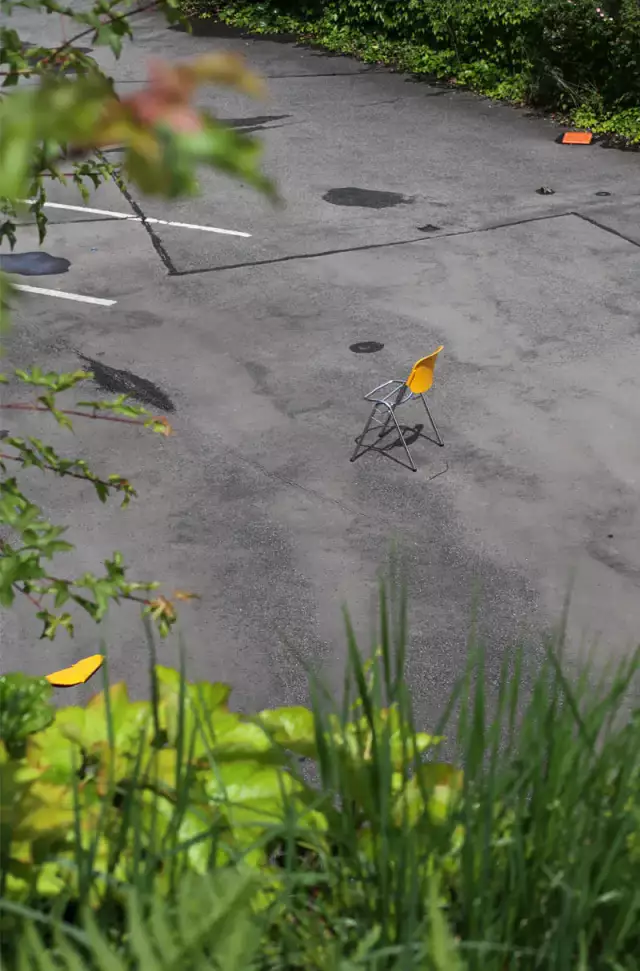 as Bild zeigt einen Stuhl, der auf einem Gehweg steht, in einer sommerlichen Atmosphäre. Die Außenumgebung umfasst Pflanzen, Gras und eine Straße im Hintergrund