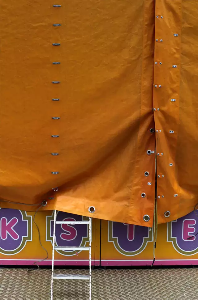 Das Foto zeigt einen orangen Plastikvorhang. Darunter schaut ein Teil von einer Leiter hervor. Unterhalb ist ein Sockel mit Buchstaben und ein Metallboden