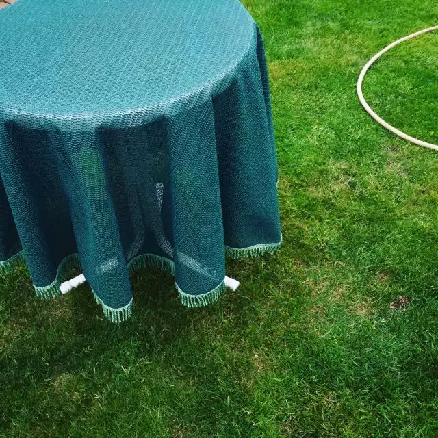 Das Foto zeigt einen runden Tisch mit grüner Tischdecke, der draußen auf Gras steht, daneben liegt ein Teil von einem Schlauch