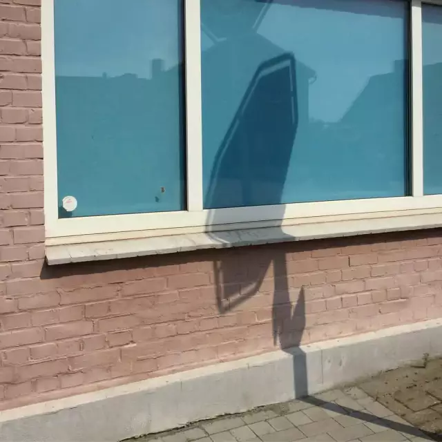 Das Foto zeigt eine rosa Hauswand mit hellblauem Fenster, in dem sich der Schatten von einem Verkehrsschild spiegelt