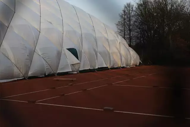 Das Foto zeigt ein großes weißes Zelt, das im Freien aufgebaut ist. Es steht auf einem roten Tennisplatz