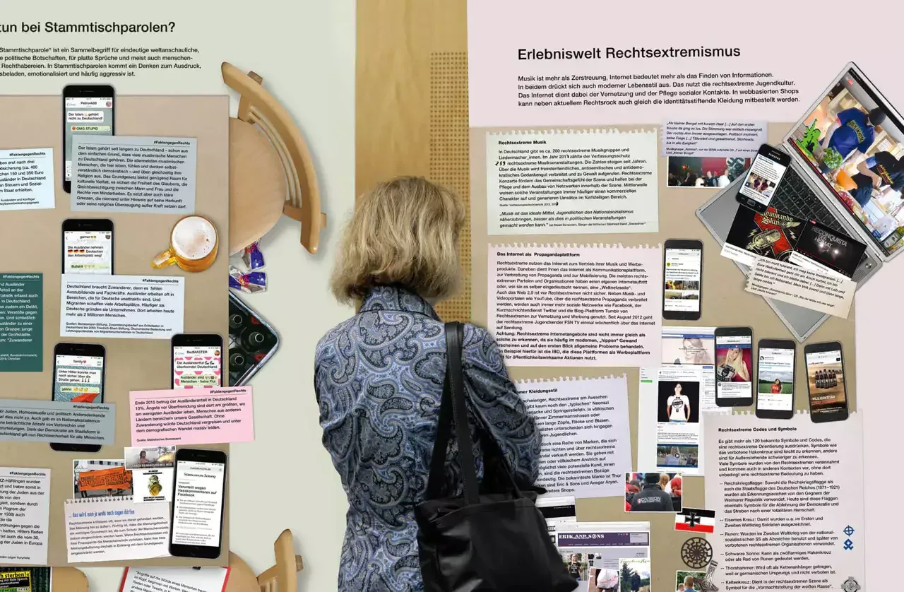 Eine Person von hinten betrachtet studiert eine großformatige Ausstellungswand mit mehreren Textblöcken, Fotografien und Informationsmaterialien zum Thema Rechtsextremismus. Die Wand ist in verschiedenen Farben gestaltet und enthält Links zu digitalen Inhalten sowie gedruckte und mobile Elemente.