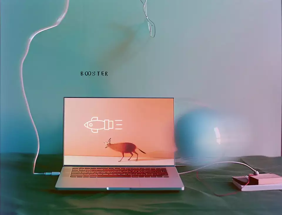 Das Bild zeigt einen Screenshot einer Website, auf der das Wort BOOSTER angezeigt wird. Weiter sind abgebildet: Computer, Laptop, merkwürdiges elektronisches Gerät und ein verschwommener Ball. Im Stil eines Stilllebens.