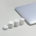 Vier weiße Behälter in aufsteigender Größe neben einem silbernen Laptop auf weißem Hintergrund. Die Behälter scheinen Pillen oder Tabletten zu enthalten und demonstrieren möglicherweise unterschiedliche Dosierungen oder Verpackungsgrößen.