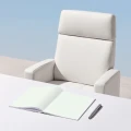 Ein weißer ergonomischer Bürostuhl mit Kopfstütze steht hinter einem weißen Schreibtisch, auf dem ein aufgeschlagenes Notizbuch und ein Stift liegen. Die Szene zeigt ein minimalistisches Arbeitsplatz-Setup vor hellblauem Hintergrund.