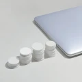 Vier weiße Behälter in aufsteigender Größe neben einem silbernen Laptop auf weißem Hintergrund. Die Behälter scheinen Pillen oder Tabletten zu enthalten und demonstrieren möglicherweise unterschiedliche Dosierungen oder Verpackungsgrößen.
