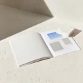 Ein offenes weißes Katalog oder Portfolio liegt auf einer hellen Betonoberfläche und zeigt auf der rechten Seite Farbmuster in Blau und Grautönen sowie leere weiße Seiten auf der linken Seite.