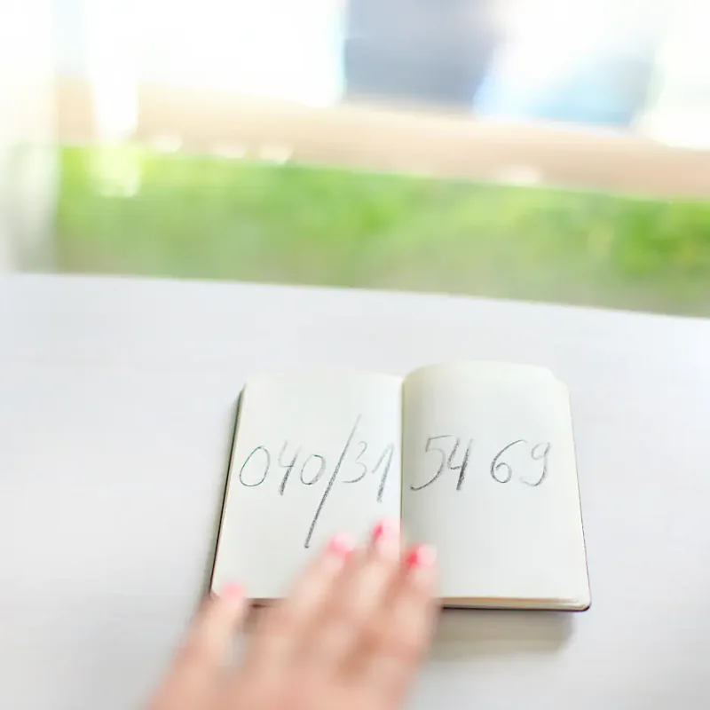 Hand zeigt auf ein aufgeschlagenes Notizbuch mit Zahlen auf einem weißen Tisch, im Hintergrund grüne Pflanzen und ein heller Raum.