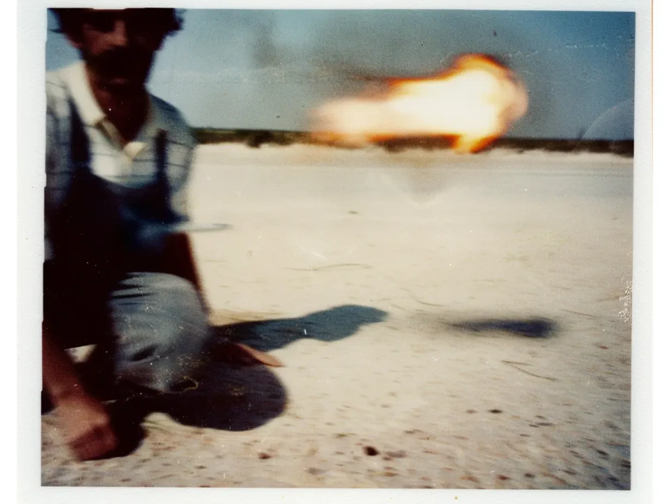 Eine Person in Jeans und Hemd kniet auf sandigem Boden und feuert eine Rakete ab, aus deren Lauf Flammen schießen. Die Aufnahme wirkt wie eine polaroidähnliche Fotografie mit warmen Farbtönen und unscharfem Hintergrund.