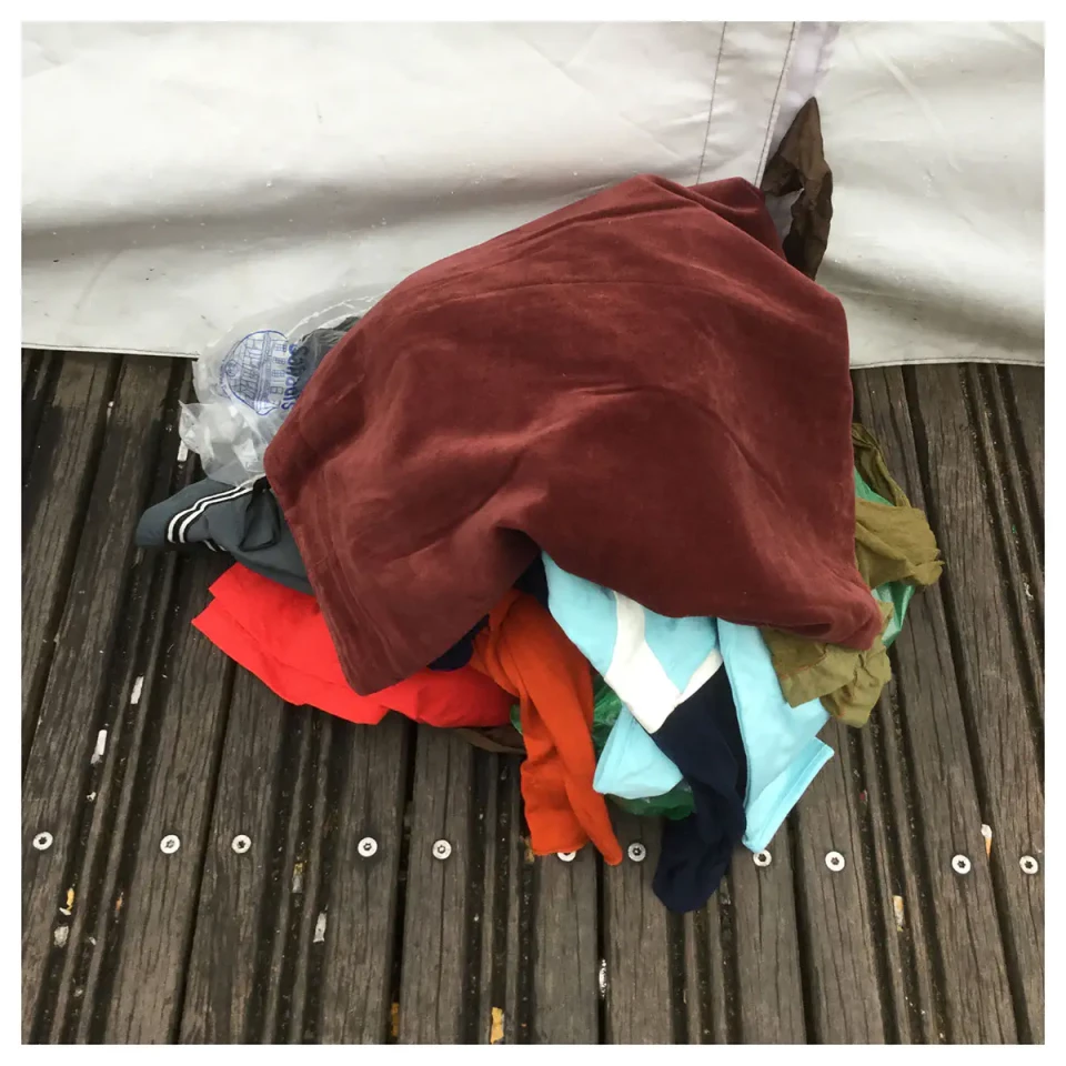 Ein Bündel bunter Stoffe in Rot, Orange, Blau und anderen Farben liegt auf einem Holzsteg. Darunter sind verschiedene textile Materialien und Fetzen zu sehen, die locker übereinander geschichtet sind.