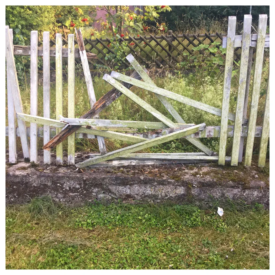 Ein weißes Holztor mit Querbalken liegt zerbrochen und zerstört im Gras, dahinter ist ein Maschendrahtzaun und grüne Vegetation sichtbar. Die beschädigte Struktur symbolisiert Barrieren und Trennung.