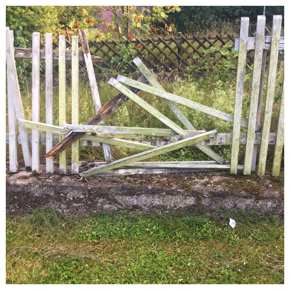 Ein weißes Holztor mit Querbalken liegt zerbrochen und zerstört im Gras, dahinter ist ein Maschendrahtzaun und grüne Vegetation sichtbar. Die beschädigte Struktur symbolisiert Barrieren und Trennung.