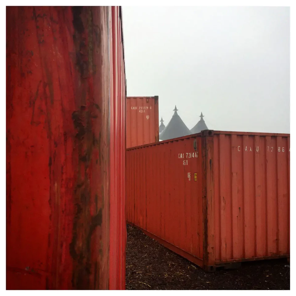 3 rote Container stufig nebeneinader stehend, im Hintergrund 2 Zeltspitzen, die wie Kirchtürme wirken