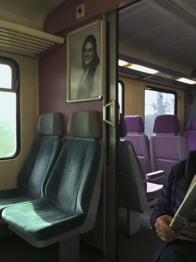Interior eines Zugs mit grauen und violetten Polstersitzen, geöffneter Tür und einem Portrait an der Wand.