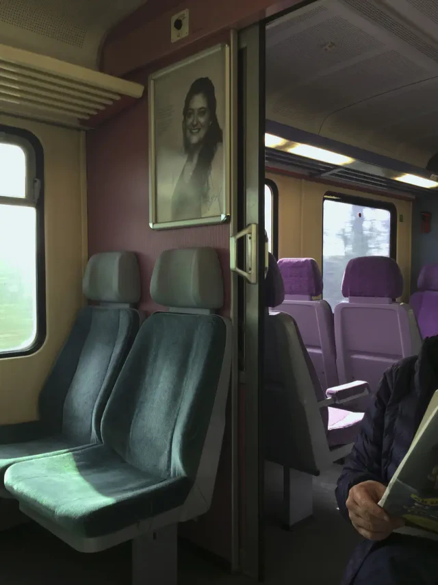 Interior eines Zugs mit grauen und violetten Polstersitzen, geöffneter Tür und einem Portrait an der Wand.