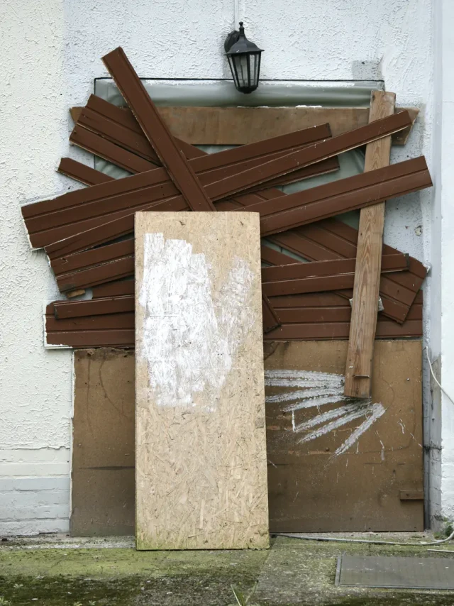 Eine große hölzerne Tür ist mit gekreuzten braunen Holzbrettern blockiert, die ein X-Muster bilden. Eine hellbraune Sperrholzplatte lehnt davor. Die Tür befindet sich an einer weißen Wand mit einer schwarzen Laterne oben.