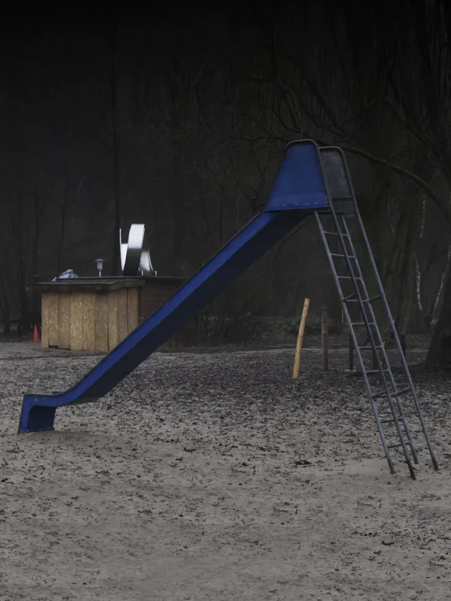 Ein blauer Spielplatz-Rutsch mit Metallleiter auf sandigem Untergrund, daneben eine hölzerne Box-Struktur vor dunklem Hintergrund.