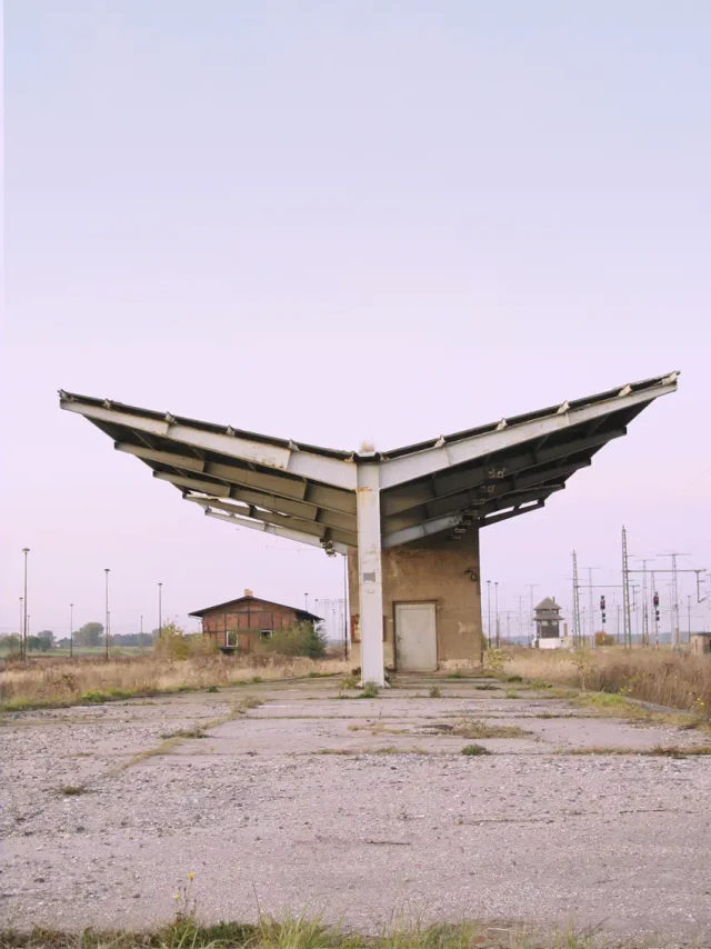 Ein verlassenes, symmetrisches Dachkonstrukt mit zwei nach oben zeigenden Flügeln steht auf einem Betonplatz in einer öden Industrielandschaft. Eine kleine Holzhütte und verschiedene Strommasten sind im Hintergrund sichtbar. Die Szenerie wirkt wie ein verfallener, surrealer Ort an einem verlassenen Industriegelände.