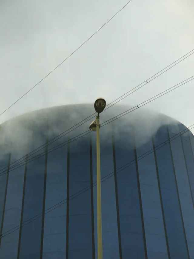 Gelber Strommast mit mehreren Stromleitungen vor einem großen, nebelverhüllten Kuppeldach aus vertikalen blauen Lamellen. Der Himmel ist grau und bewölkt.