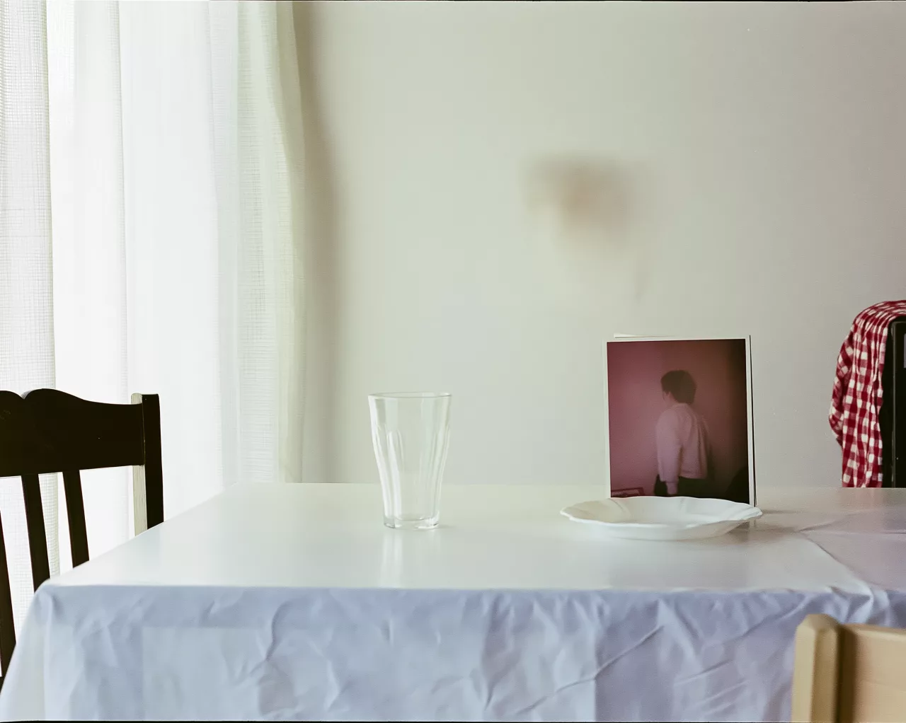 Minimalistische Szene mit Glas und Teller auf weißem Tisch, unscharfem Schatten an der Wand und teilweise sichtbarer Person am rechten Bildrand.