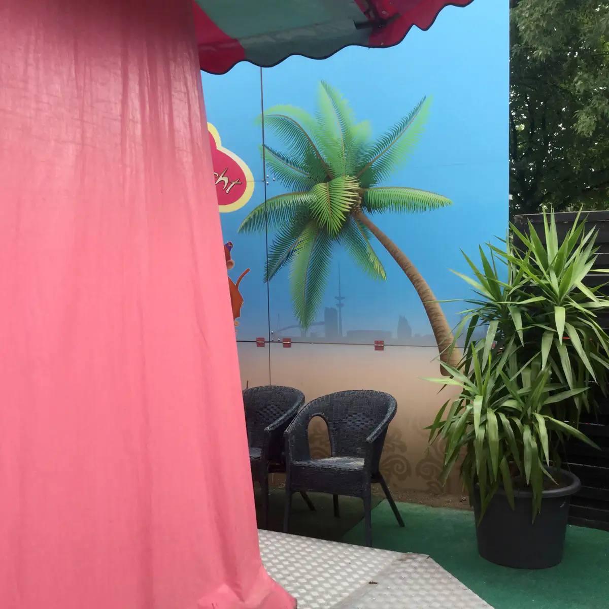 Ein bunter Innenraum mit rosa Vorhang, einer gemalten Strandszene mit Palme an der Wand, schwarzen Sesseln und grünen Pflanzen in Töpfen. Der Raum wirkt wie ein entspannendes Paradies-Thema-Setting.