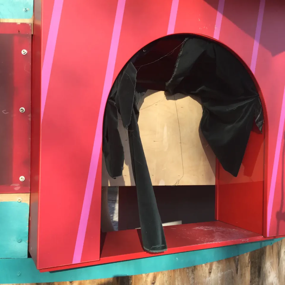 Ein schwarzer Blazer hängt auf einem Kleiderbügel in einer roten Nische mit türkisfarbenen Akzenten. Der Blazer wird vor einem beigen Hintergrund präsentiert. Die Ausstellung ist Teil der Kollektion "Paradies" auf IconScreen.