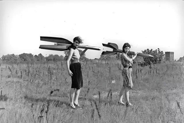 Zwei junge Frauen - Raketenmädchen - tragen Holzraketen auf ihren Schultern und wenden den Blick auf den Betrachter.