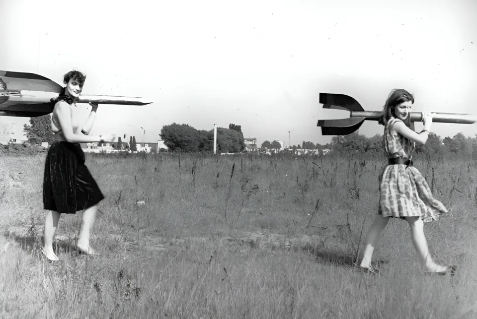 Zwei junge Frauen - Raketenmädchen - tragen Holzraketen auf ihren Schultern. Im Hintergrund eine karge Landschaft.