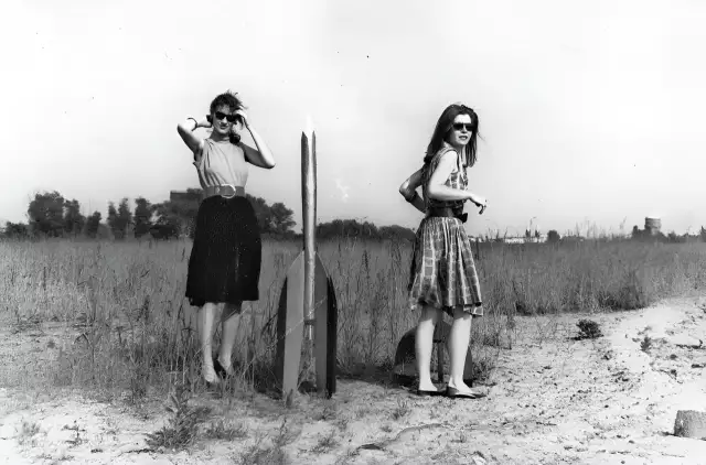 Zwei junge Frauen - Raketenmädchen - stehen neben einer Holzrakete. Die schwarz-weiße Aufnahme zeigt sie in Kleidern und mit einer Sonnenbrille.