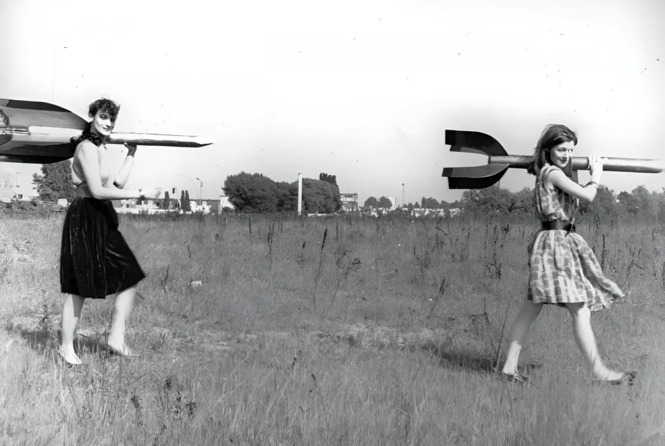 Zwei junge Frauen - Raketenmädchen - tragen Holzraketen auf ihren Schultern. Im Hintergrund eine karge Landschaft.