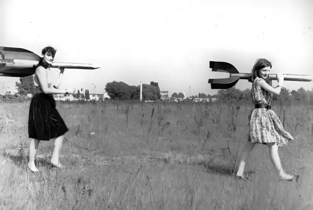 Zwei junge Frauen - Raketenmädchen - tragen Holzraketen auf ihren Schultern. Im Hintergrund eine karge Landschaft.