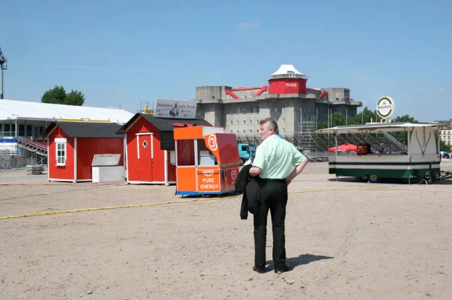 Ein Mann in grünem Hemd und dunkler Hose steht auf einem asphaltierten Platz vor roten und orangefarbenen Holzbauten, darunter ein Schaustellerstand. Im Hintergrund sind weitere Gebäude und ein Turm unter blauem Himmel sichtbar.
