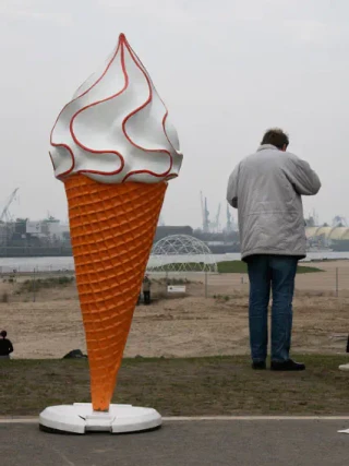Ein übergroßes Eiscreme-Kunstwerk mit Waffelkegel und weißer Sahne-Haube steht auf einem freien Platz, während eine Person in grauer Jacke davor steht und es betrachtet. Im Hintergrund sind Industrieanlagen und Wasser sichtbar.