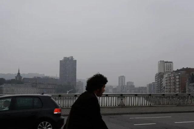 Eine Person in schwarzer Kleidung geht über eine Brücke mit einer grauen Stadtlandschaft im Hintergrund. Ein Auto ist links zu sehen, während mehrstöckige Gebäude durch den Nebel verschwommen sind.