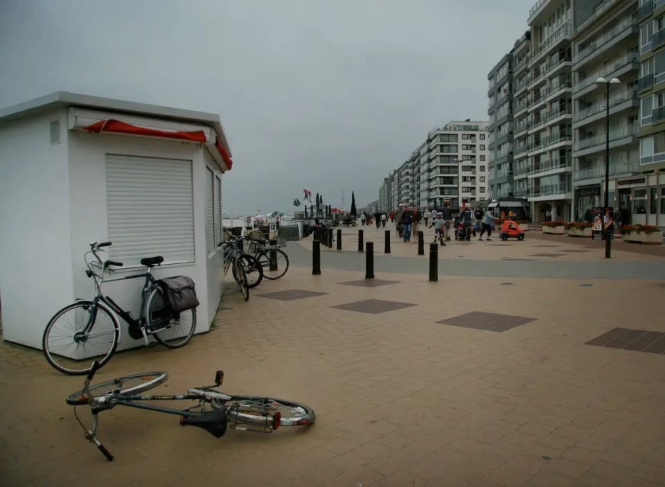 Ein Fahrrad liegt auf einem breiten Strand vor einer weißen Umkleidekabine und Fahrradständern. Im Hintergrund erstreckt sich ein Promenadenpfad mit Pollern und Hochhäusern entlang der Küste. Der Himmel ist bewölkt und der Strand wirkt verlassen.