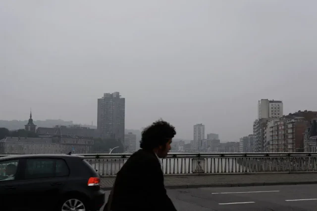 Eine Person in schwarzer Kleidung geht über eine Brücke mit einer grauen Stadtlandschaft im Hintergrund. Ein Auto ist links zu sehen, während mehrstöckige Gebäude durch den Nebel verschwommen sind.