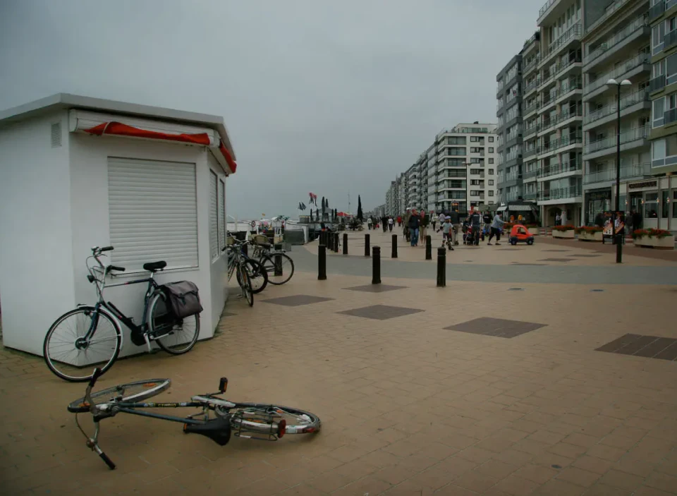 Ein Fahrrad liegt auf einem breiten Strand vor einer weißen Umkleidekabine und Fahrradständern. Im Hintergrund erstreckt sich ein Promenadenpfad mit Pollern und Hochhäusern entlang der Küste. Der Himmel ist bewölkt und der Strand wirkt verlassen.
