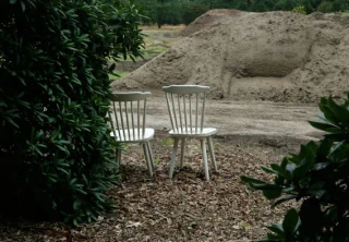 Zwei weiße Stühle stehen auf einem Kiesplatz zwischen grünen Büschen, mit Sandhaufen im unscharfen Hintergrund. Die minimalistische Anordnung wirkt wie eine meditative oder künstlerische Installation in einer Gartenlandschaft.