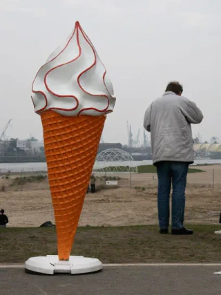 Ein übergroßes Eiscreme-Kunstwerk mit Waffelkegel und weißer Sahne-Haube steht auf einem freien Platz, während eine Person in grauer Jacke davor steht und es betrachtet. Im Hintergrund sind Industrieanlagen und Wasser sichtbar.