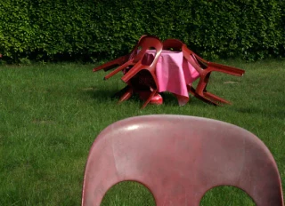 Ein großes rotes Kunstwerk in Form eines Käfers mit ausgebreiteten Flügeln steht auf einer grünen Wiese vor einer Hecke. Im Vordergrund ist eine rote, glänzende Maske mit zwei großen ovalen Öffnungen für die Augen sichtbar.