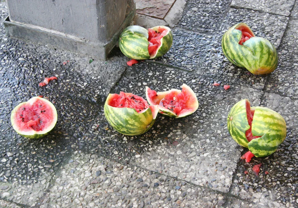 Mehrere aufgeschnittene Wassermelonen liegen auf einem grauen Pflasterbelag. Die Melonen zeigen ihr rotes Fruchtfleisch und grüne Schalen, einige sind halbiert, andere in Stücke geschnitten.
