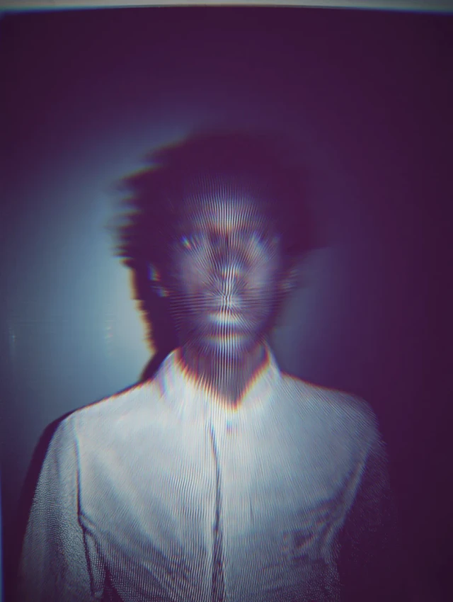 Person mit verschwommenem Gesicht trägt weißes Hemd vor dunklem Hintergrund. Das Porträt hat einen glitchartigen, pixelierten Effekt mit roten und blauen Farbversätzen. Die Aufnahme wirkt experimentell und digital verzerrt.