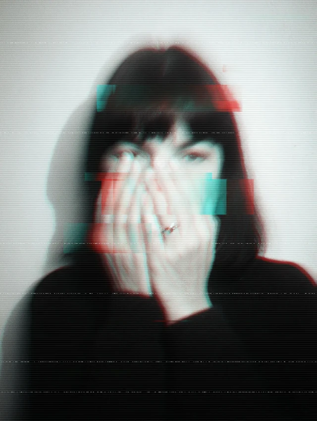 Person mit schwarzem Haar und Pony hält eine Hand vor das Gesicht, aufgenommen mit Glitch-Effekt in Rot und Türkis.