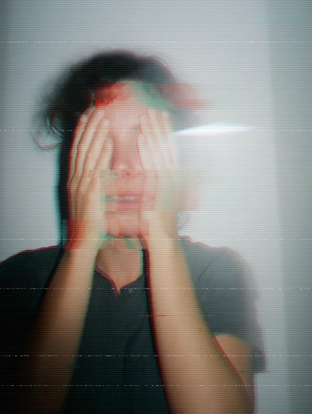 Person mit gesenktem Kopf, Gesicht mit beiden Händen verborgen, trägt schwarzes Shirt, mit Chromatic-Aberration-Effekt und Scan-Linien-Artefakten.