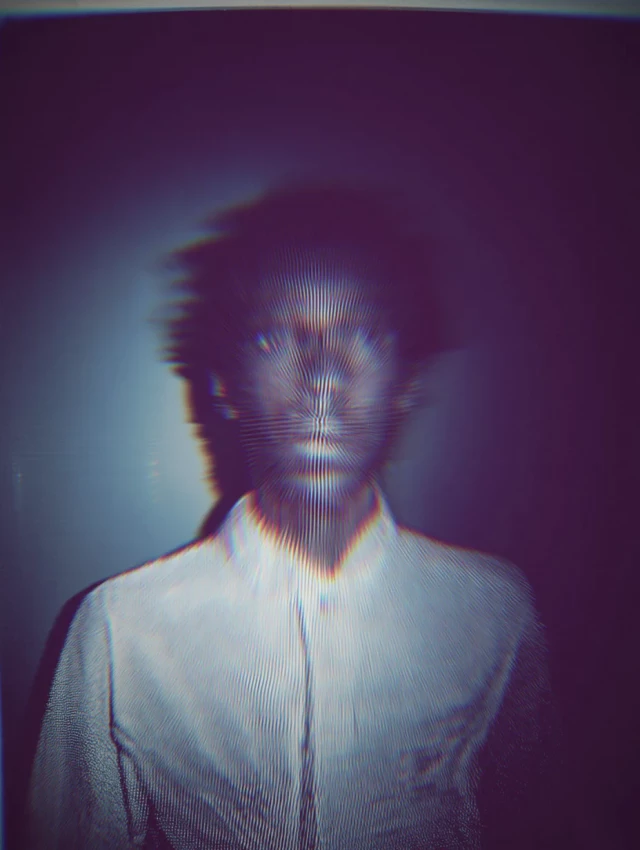 Person mit verschwommenem Gesicht trägt weißes Hemd vor dunklem Hintergrund. Das Porträt hat einen glitchartigen, pixelierten Effekt mit roten und blauen Farbversätzen. Die Aufnahme wirkt experimentell und digital verzerrt.