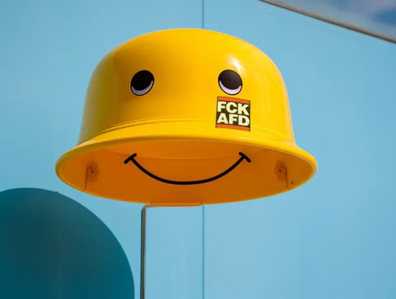 Gelber Helm mit lächelndem Gesicht und der Aufschrift "FCK AFD" vor blauem Himmel.