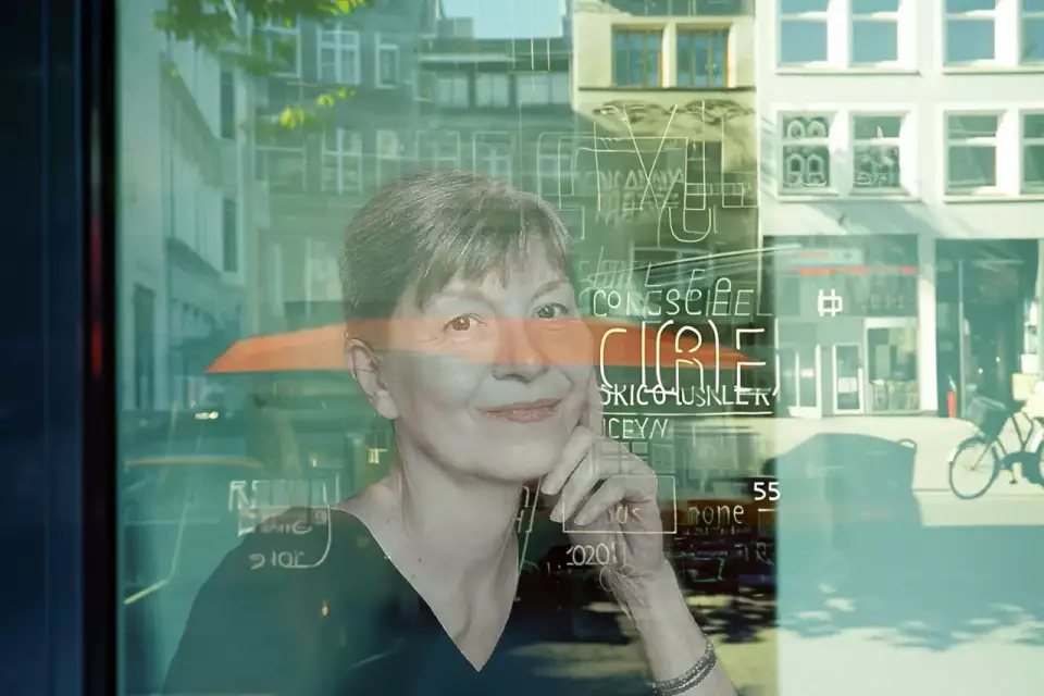 Das Bild zeigt Ingrid Völlmeke mit der Hand am Gesicht. Der Inhalt umfasst den Text ICONSCREEN. Stichwörter die das Bild beschreiben: menschliches Gesicht, Gebäude, Person, Lächeln, Fenster, im Freien und Reflexion.