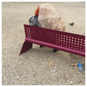 eine rothaarige Frau sitz auf einer Bank im Park und schütz sich gegen den Wind mit einem Regenschirm, der sich mit einem ähnlichen Muster wie der Schotter unter der Bank darstellt, skurril